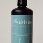 Bottle of 'Tan Alchemy' product on a reflective surface with a neutral background