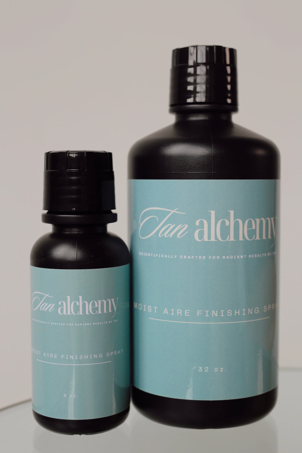 Two bottles of 'Tan alchemy' on a reflective surface with a neutral background