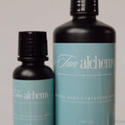 Two bottles of 'Tan alchemy' on a reflective surface with a neutral background