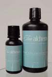 Two bottles of 'Tan alchemy' on a reflective surface with a neutral background