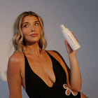 Woman in a black swimsuit holding a Self Tanning Mousse - Tampa Bay Tan against a plain background