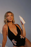 Woman in a black swimsuit holding a Self Tanning Mousse - Tampa Bay Tan against a plain background