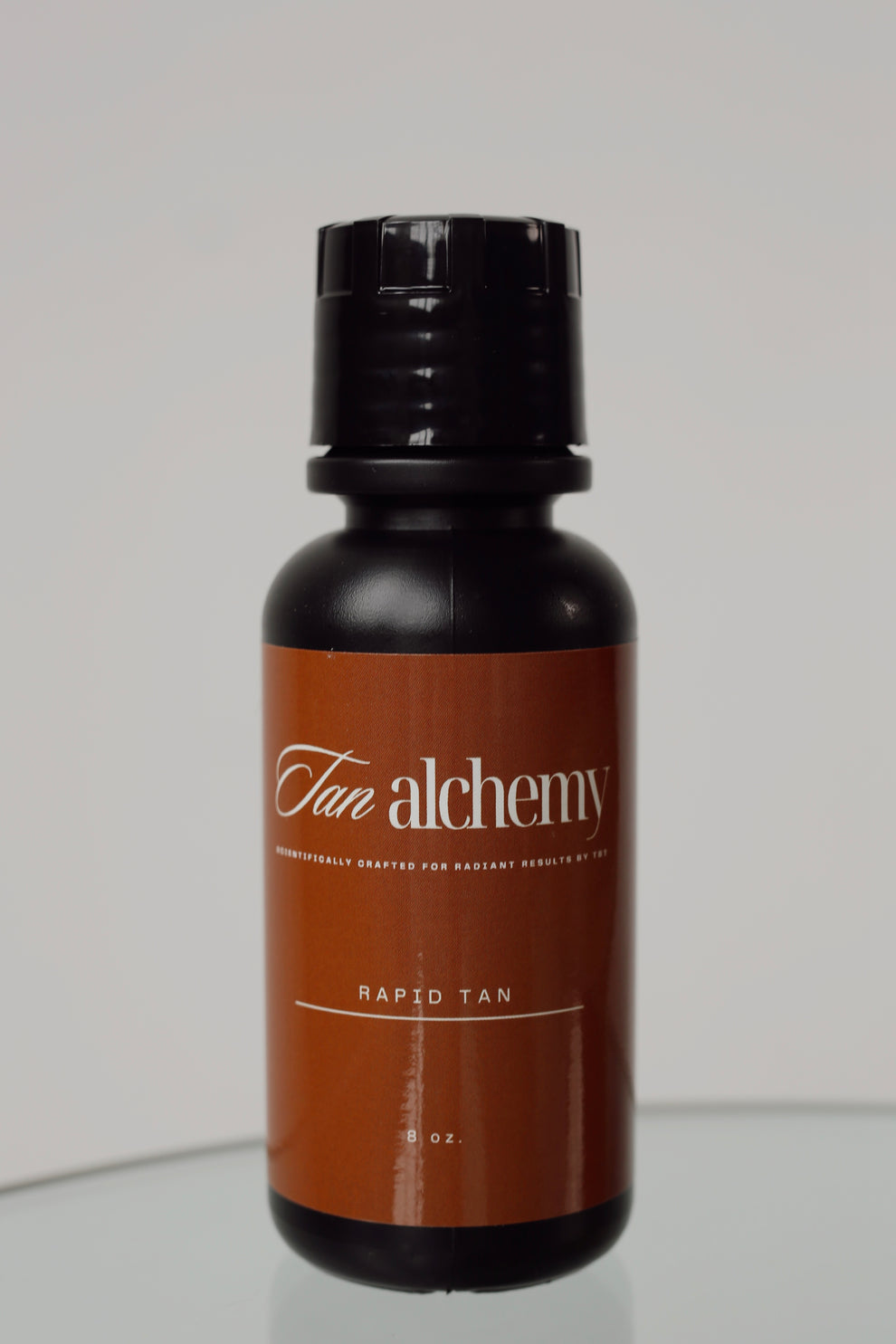 Brown bottle with black cap labeled 'Tan Alchemy' on a light gray background