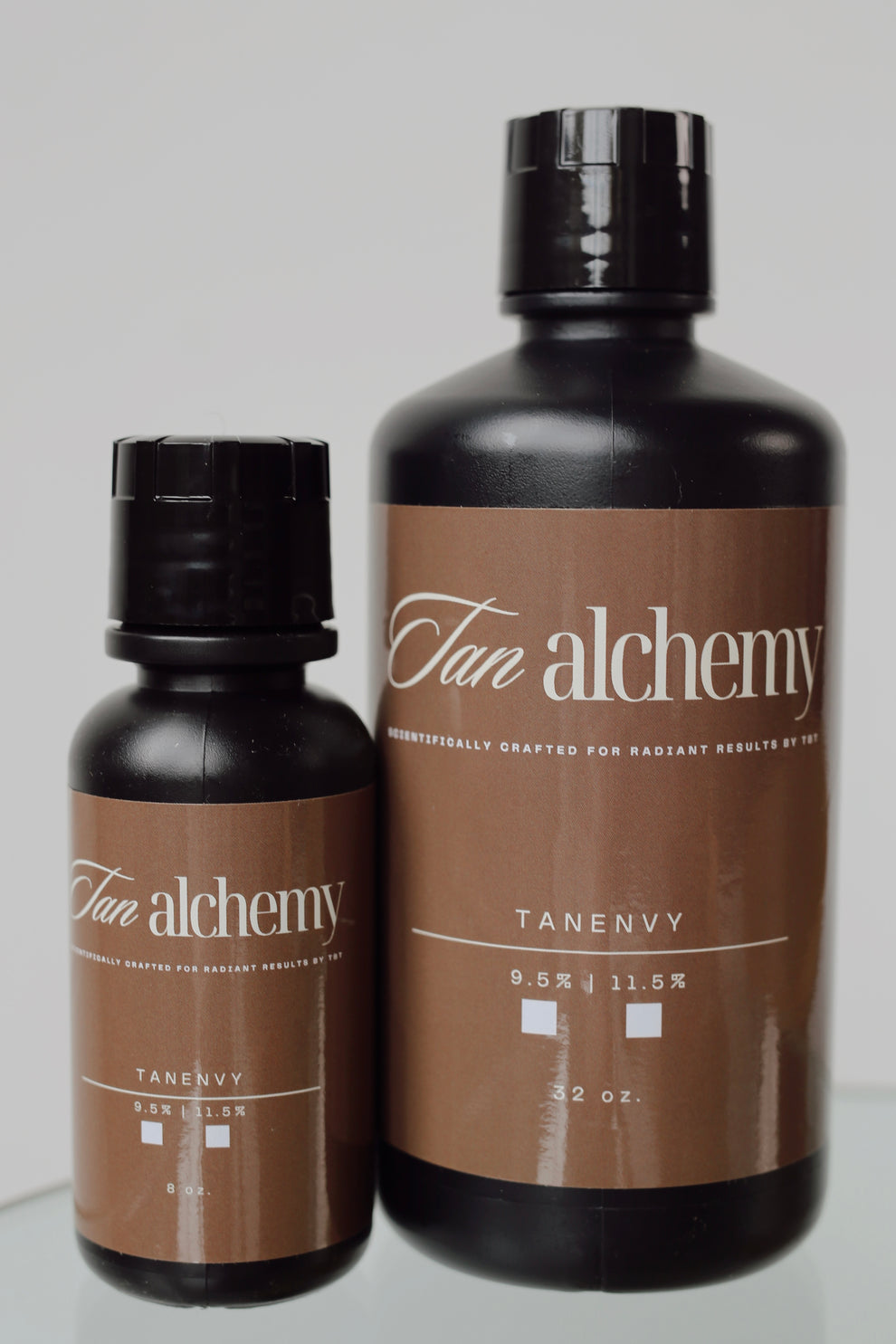 Two bottles of Tan Alchemy Tan Envy on a reflective surface with a neutral background