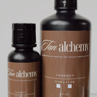 Two bottles of Tan Alchemy Tan Envy on a reflective surface with a neutral background