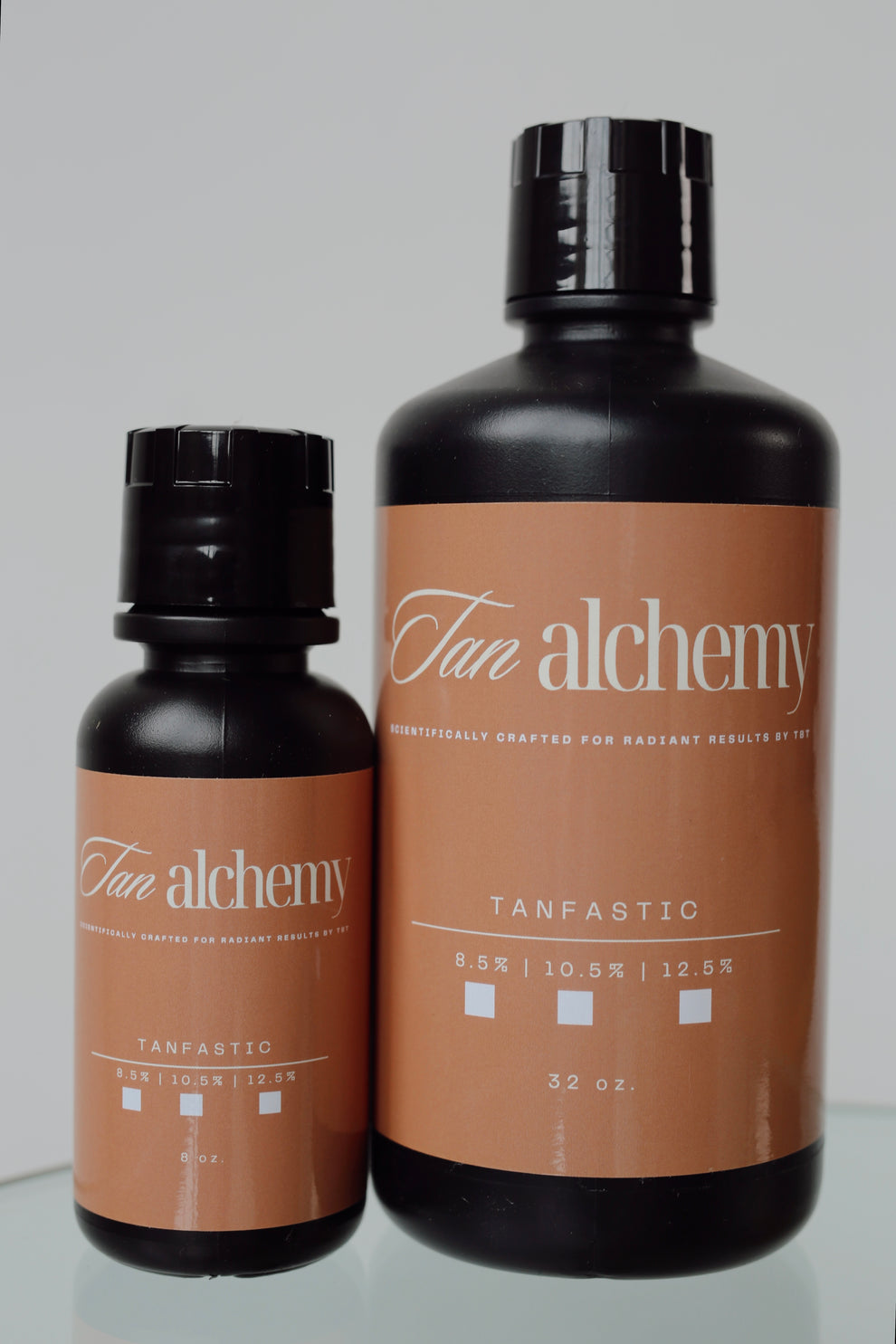Two bottles of Tan Alchemy Tan Fantastic tanning lotion on a reflective surface with a gray background.