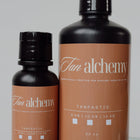 Two bottles of Tan Alchemy Tan Fantastic tanning lotion on a reflective surface with a gray background.