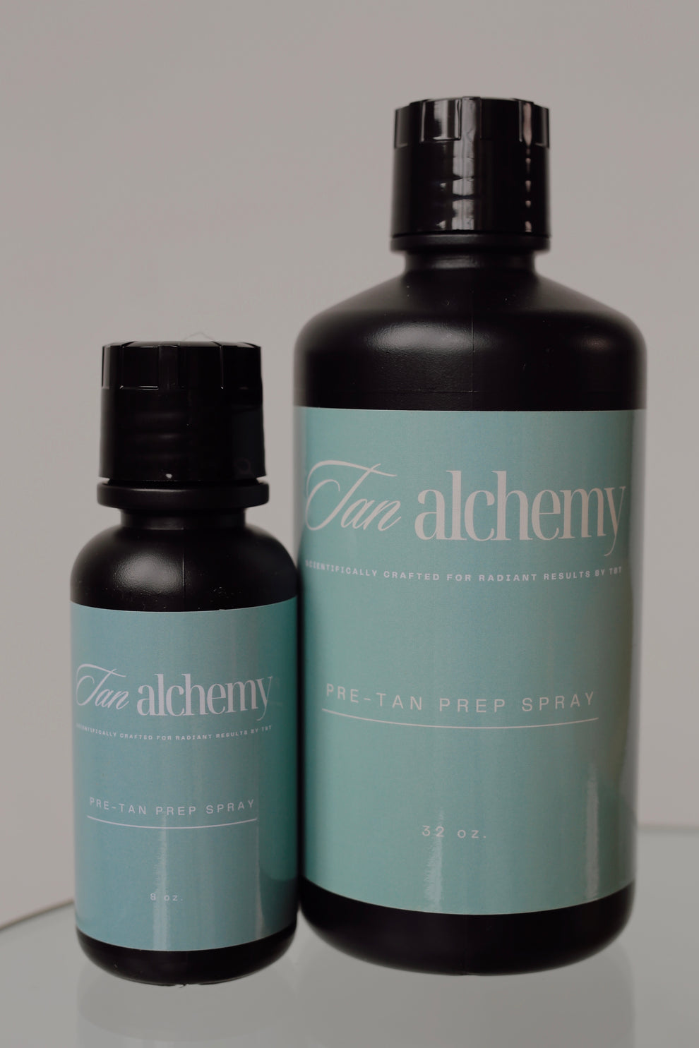 Two bottles of Tan Alchemy pre-tan prep spray on a gray background