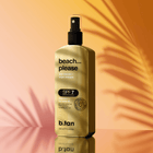Gold bottle of b.tan 'beach... please' sunscreen spray on a warm gradient background