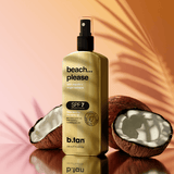 Gold bottle of b.tan 'beach... please' sunscreen spray with coconuts on a reflective surface.