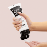 Hand holding a tube of 'erase tan repeat' product against a beige background