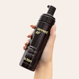 Hand holding a black bottle labeled 'tanned AF' with 'b.tan' branding on a beige background