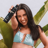 Woman in a bikini holding a bottle of 'b.tan' tanning lotion with green leaves in the background