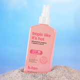 Pink bottle of b.tan sun spray with 'tropic like it's hot' text on a sandy surface with a blue gradient background