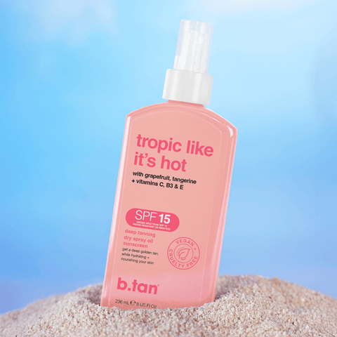 Pink bottle of b.tan sun spray with 'tropic like it's hot' text on a sandy surface with a blue gradient background